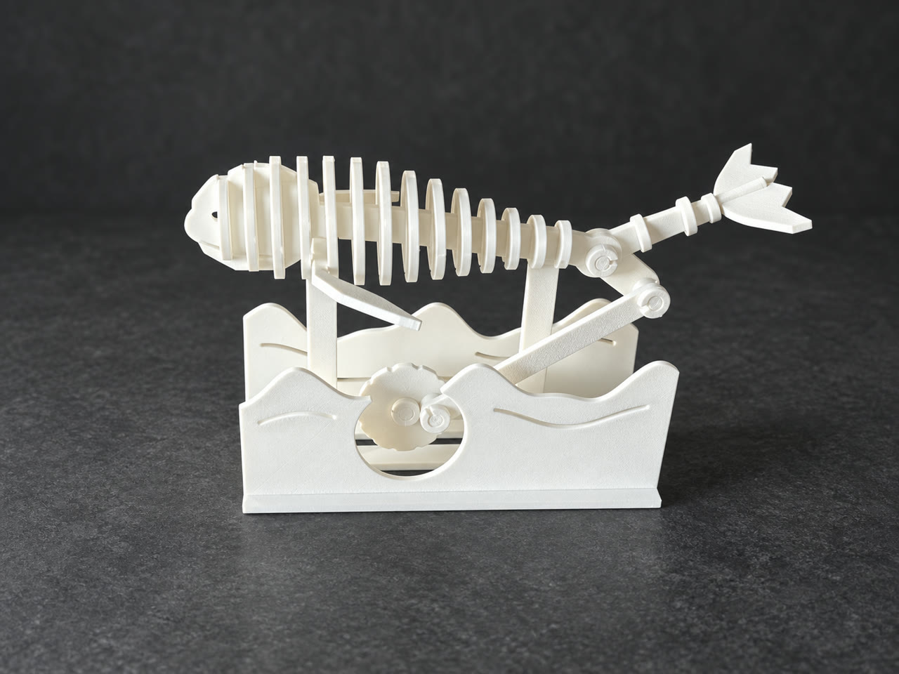White skeletal whale prototype with a crank and wave-shaped supports, presented against a retouched charcoal background