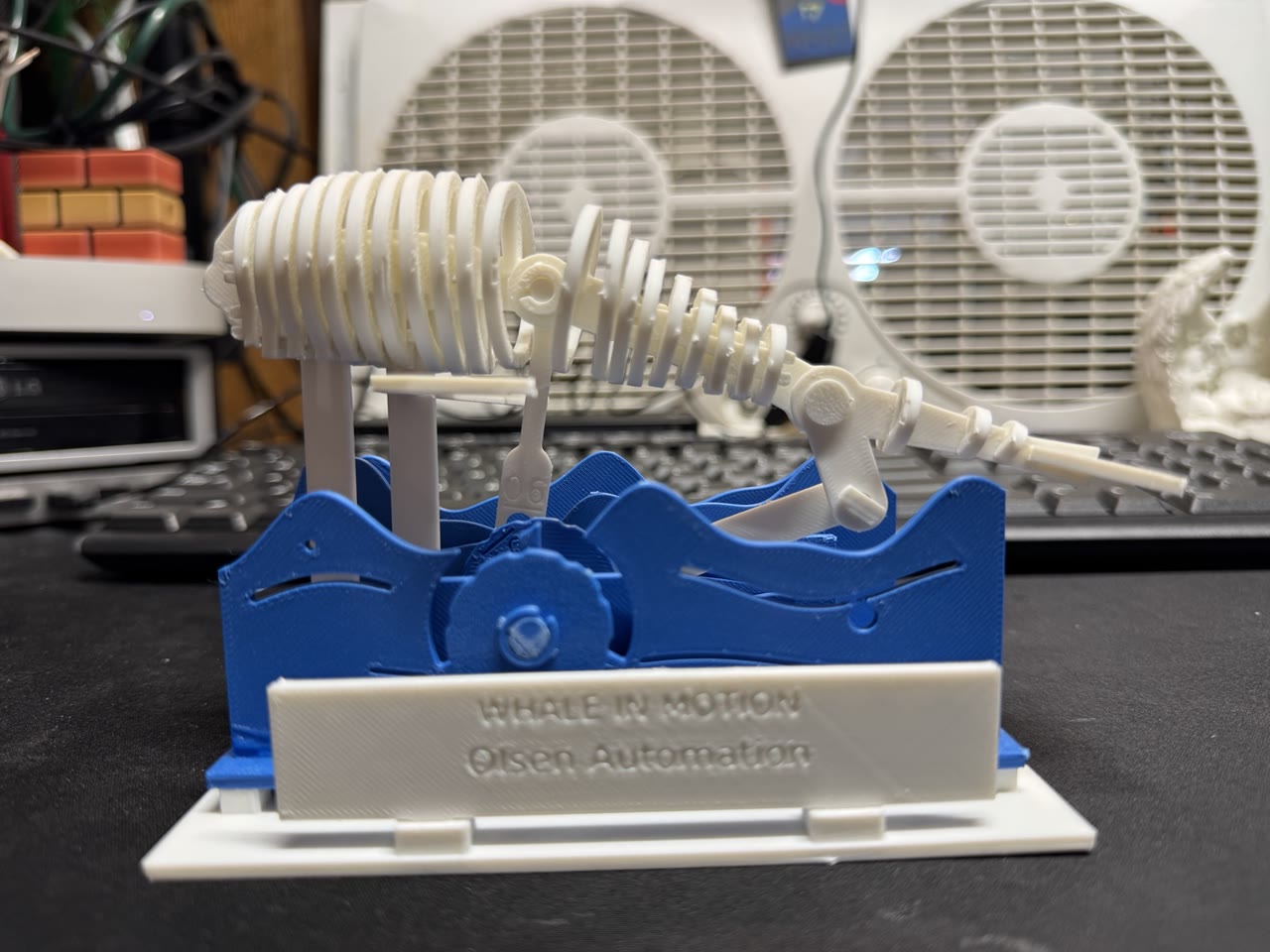 White mechanical whale over blue waves on a white platform labeled Whale in Motion and Olsen Automation, photographed on the workbench